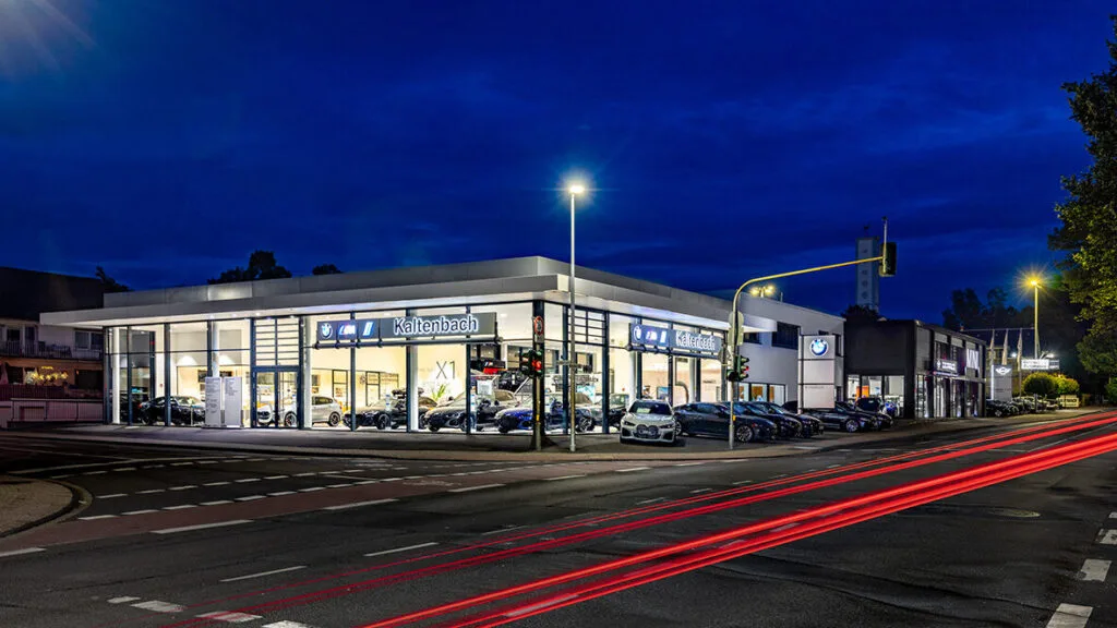 Kaltenbach Gruppe – Ein modernes Autohaus mit großen Glasfenstern ist nachts hell erleuchtet. Mehrere Autos sind innerhalb und außerhalb des Gebäudes ausgestellt. Auf der Straße davor sind rote Streifen von den Scheinwerfern vorbeifahrender Autos zu sehen. – BMW, MINI, KIA, BMW Motorrad & Honda Motorrad Partner in NRW
