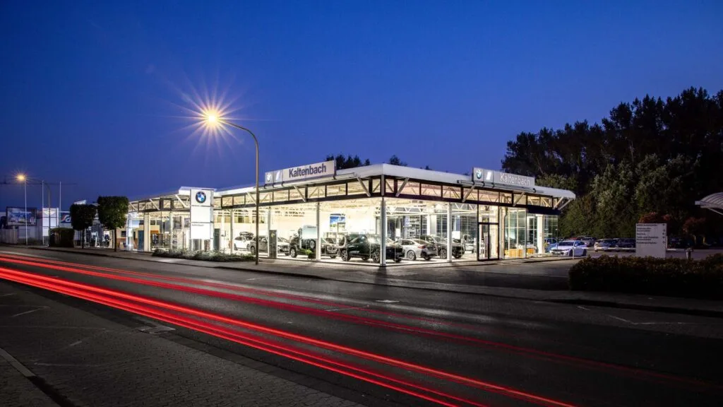 Kaltenbach Gruppe – Ein Autohaus mit Glaswänden wird in der Abenddämmerung beleuchtet, wobei mehrere Autos im Inneren zu sehen sind. Auf der Straße davor sind rote Lichtspuren von vorbeifahrenden Fahrzeugen zu sehen. Bäume säumen den Hintergrund, und Straßenlaternen beleuchten die Szene. – BMW, MINI, KIA, BMW Motorrad & Honda Motorrad Partner in NRW