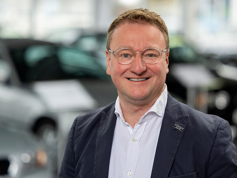Kaltenbach Gruppe – Ein Mann mit kurzen hellbraunen Haaren, der eine Brille, ein weißes Hemd und einen marineblauen Blazer trägt, lächelt in die Kamera. Im Hintergrund sind unscharfe Autos zu sehen, die auf einen Ausstellungsraum oder ein Autohaus hindeuten. – BMW, MINI, KIA, BMW Motorrad & Honda Motorrad Partner in NRW