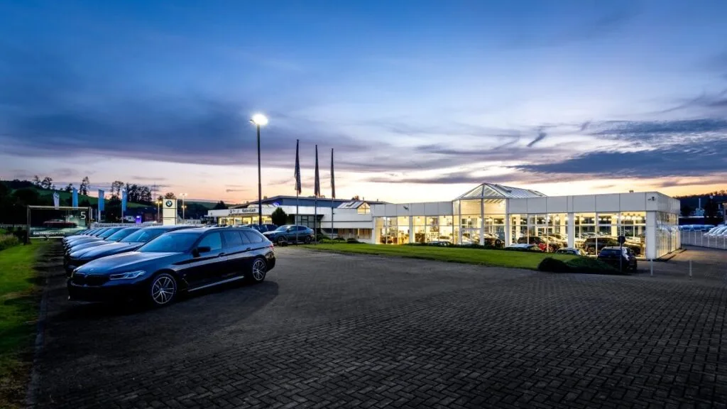 Kaltenbach Gruppe – Eine Reihe von Autos ist vor einem hell erleuchteten Autohaus in der Abenddämmerung geparkt, mit einem klaren Himmel und weichen Wolken im Hintergrund. Die Glasfenster des Autohauses geben den Blick auf weitere Autos im Inneren frei. – BMW, MINI, KIA, BMW Motorrad & Honda Motorrad Partner in NRW