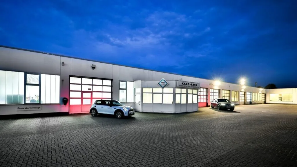 Kaltenbach Gruppe – Eine moderne Autowerkstatt mit großen gläsernen Garagentoren, die in der Abenddämmerung von innen beleuchtet werden. Zwei Autos sind vor dem Gebäude geparkt, und der Himmel ist tiefblau mit vereinzelten Wolken. – BMW, MINI, KIA, BMW Motorrad & Honda Motorrad Partner in NRW