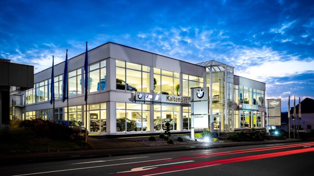Kaltenbach Gruppe – Modernes BMW-Händlergebäude in der Abenddämmerung, hell erleuchtet mit großen Glasfenstern, in denen Autos ausgestellt sind. Der Himmel ist blau und wolkenverhangen, und auf der Straße davor sind die Lichtspuren der vorbeifahrenden Autos zu sehen. – BMW, MINI, KIA, BMW Motorrad & Honda Motorrad Partner in NRW