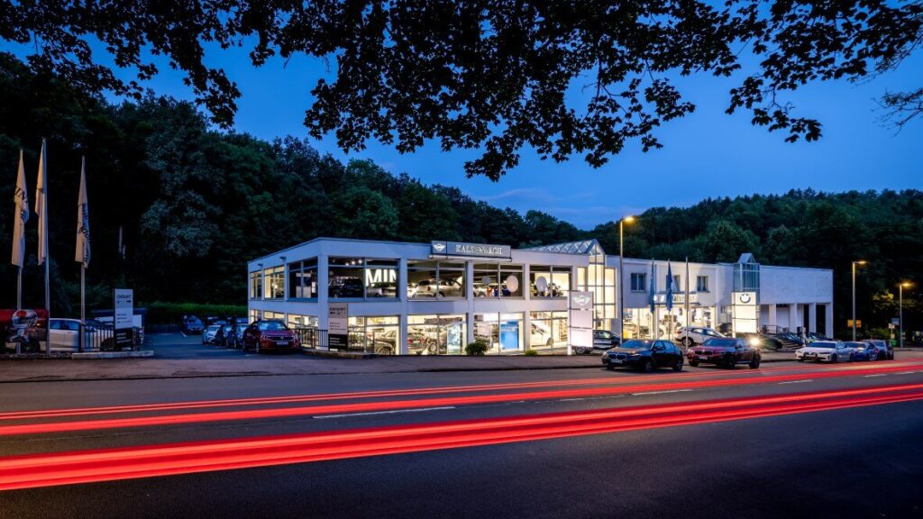 Kaltenbach Gruppe – Ein modernes Autohaus mit heller Innenbeleuchtung, großen Fenstern und mehreren draußen geparkten Autos in der Abenddämmerung. Auf der Straße davor sind rote Lichtspuren von vorbeifahrenden Fahrzeugen zu sehen. Das Gelände ist von Bäumen umgeben. – BMW, MINI, KIA, BMW Motorrad & Honda Motorrad Partner in NRW