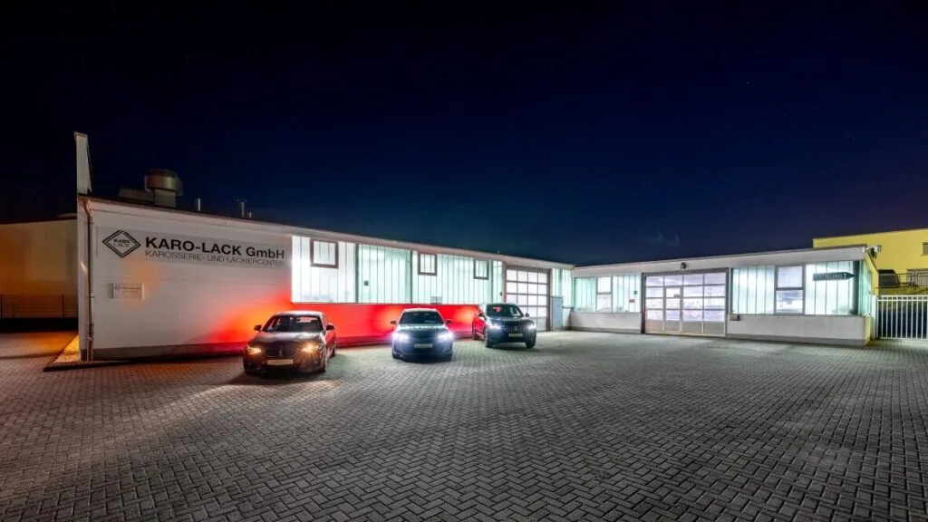 Kaltenbach Gruppe – Ein hell erleuchtetes Industriegebäude in Meinerzhagen bei Nacht, vor dem drei Autos parken. Auf dem Schild steht KARO-LACK GmbH Karosserie- und Lackierzentrum. Die Fläche ist mit Falzziegeln gepflastert. – BMW, MINI, KIA, BMW Motorrad & Honda Motorrad Partner in NRW