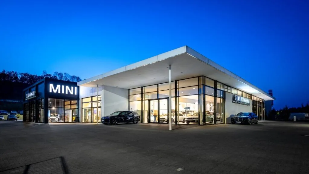 Kaltenbach Gruppe – Das Gebäude eines modernen MINI-Autohauses ist in der Abenddämmerung hell erleuchtet, mit großen Glasfenstern, in denen Autos ausgestellt sind. Der Himmel ist tiefblau und ein paar Fahrzeuge sind draußen auf einem großen Parkplatz geparkt. – BMW, MINI, KIA, BMW Motorrad & Honda Motorrad Partner in NRW