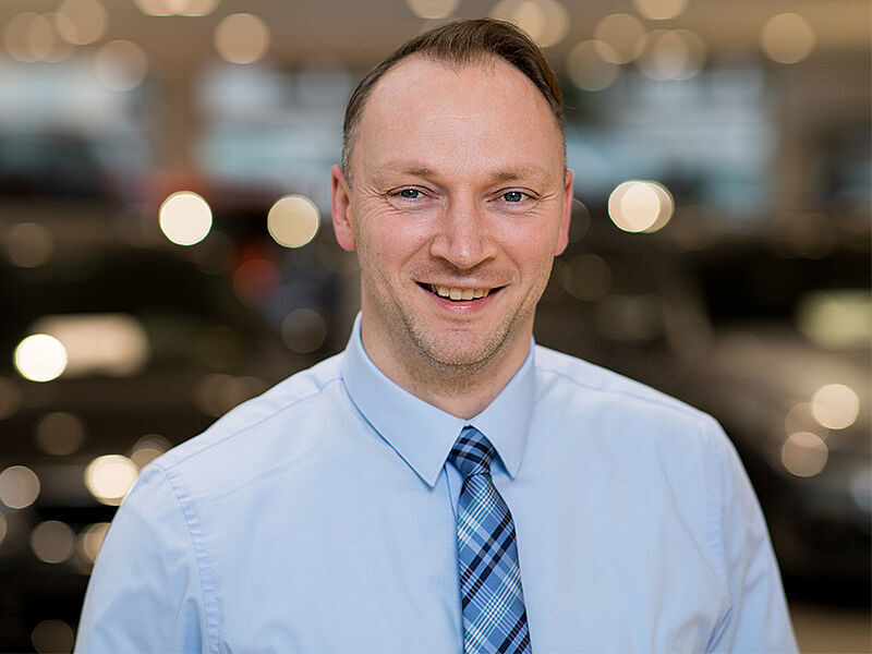 Kaltenbach Gruppe – Ein Mann mit einem hellblauen Hemd und einer blau karierten Krawatte lächelt in die Kamera. Der verschwommene Hintergrund mit kreisförmigen Lichtern schafft eine professionelle, warme Atmosphäre in Kaltenbach Lüdenscheid. – BMW, MINI, KIA, BMW Motorrad & Honda Motorrad Partner in NRW