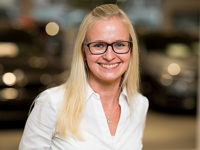 Kaltenbach Gruppe – Eine Frau mit langen blonden Haaren und Brille lächelt in die Kamera. Sie trägt eine weiße Bluse und eine Halskette. Der sanft verschwommene Hintergrund deutet die undeutlichen Lichter und dunklen Formen von Kaltenbach Lüdenscheid an. – BMW, MINI, KIA, BMW Motorrad & Honda Motorrad Partner in NRW