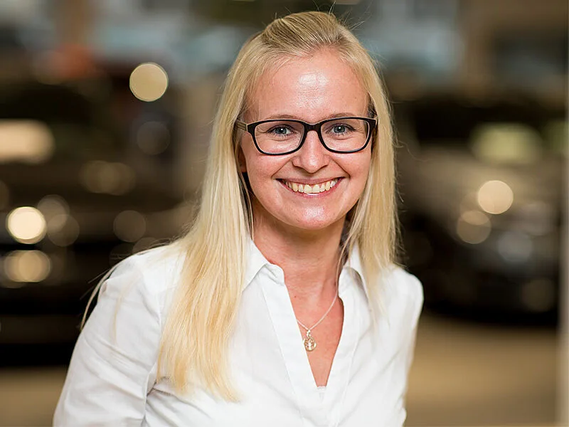 Kaltenbach Gruppe – Eine Frau mit langen blonden Haaren und Brille lächelt in die Kamera. Sie trägt eine weiße Bluse und eine Halskette. Der sanft verschwommene Hintergrund deutet die undeutlichen Lichter und dunklen Formen von Kaltenbach Lüdenscheid an. – BMW, MINI, KIA, BMW Motorrad & Honda Motorrad Partner in NRW