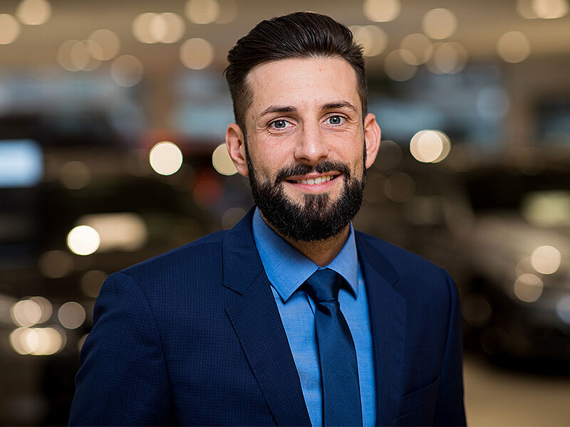 Kaltenbach Gruppe – Ein Mann mit dunklen Haaren und einem Bart, der einen blauen Anzug, ein hellblaues Hemd und eine blaue Krawatte trägt, lächelt in die Kamera. Der unscharfe Hintergrund mit warmen Lichtern deutet auf eine Veranstaltung in Kaltenbach Lüdenscheid hin. – BMW, MINI, KIA, BMW Motorrad & Honda Motorrad Partner in NRW
