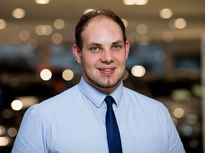 Kaltenbach Gruppe – Ein Mann mit kurzem braunem Haar und gestutztem Bart, der ein hellblaues Hemd und eine dunkelblaue Krawatte trägt, lächelt in die Kamera. Der sanft verschwommene Hintergrund mit runden Lichtern deutet auf eine Kaltenbach-Lüdenscheider Umgebung hin. – BMW, MINI, KIA, BMW Motorrad & Honda Motorrad Partner in NRW