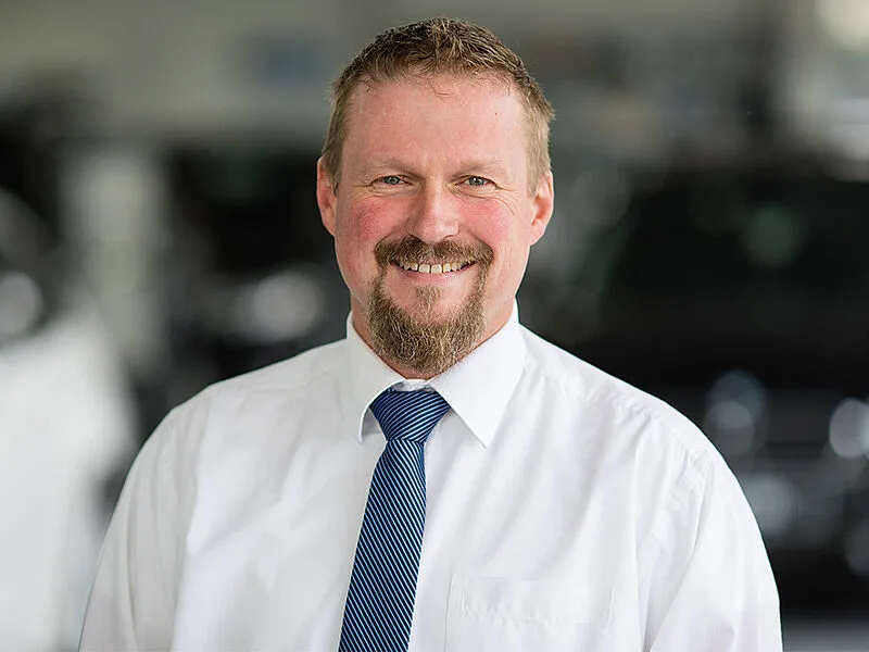 Kaltenbach Gruppe – Ein lächelnder Mann mit kurzen Haaren und Ziegenbart, weißem Hemd und blau gestreifter Krawatte, steht in einem Haus in Kaltenbach Meinerzhagen mit unscharfen Autos im Hintergrund. – BMW, MINI, KIA, BMW Motorrad & Honda Motorrad Partner in NRW