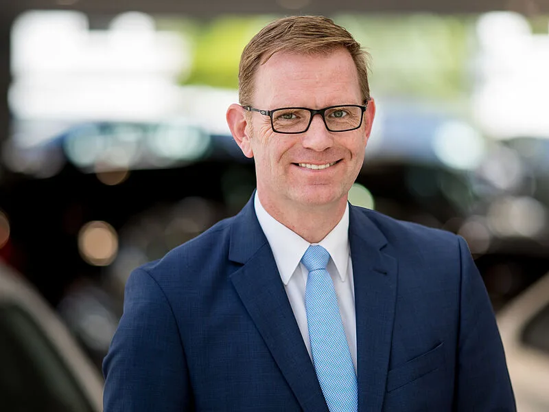 Kaltenbach Gruppe – Ein Mann in einem blauen Anzug, mit hellblauer Krawatte und Brille lächelt in die Kamera. Er hat kurzes braunes Haar und steht in einer unscharfen Innen- oder Außenumgebung mit Autos im Hintergrund. – BMW, MINI, KIA, BMW Motorrad & Honda Motorrad Partner in NRW