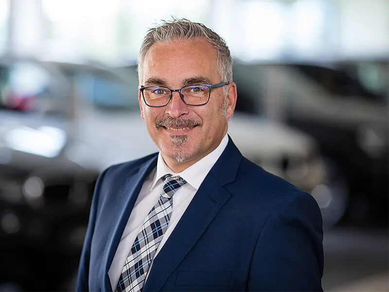 Kaltenbach Gruppe – Ein Mann mittleren Alters mit grauem Haar, Brille und Spitzbart lächelt, während er einen marineblauen Anzug, ein weißes Hemd und eine karierte Krawatte trägt. Der unscharfe Hintergrund deutet auf geparkte Autos in einem Gebäude in Kaltenbach Bergisch Gladbach hin. – BMW, MINI, KIA, BMW Motorrad & Honda Motorrad Partner in NRW