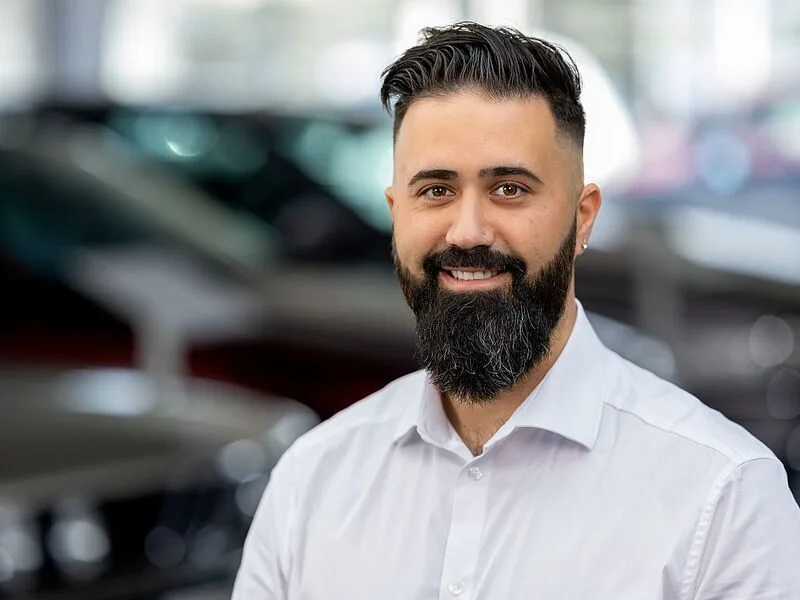 Kaltenbach Gruppe – Ein Mann mit dunklem Haar und Vollbart, der ein weißes Button-up-Hemd trägt, lächelt, während er in einem Haus in Kaltenbach-Meinerzhagen steht, mit unscharfen Autos im Hintergrund. – BMW, MINI, KIA, BMW Motorrad & Honda Motorrad Partner in NRW