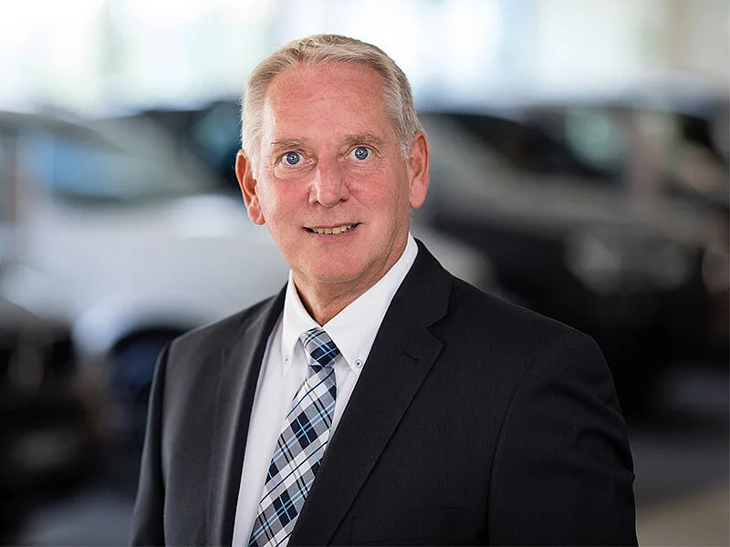 Kaltenbach Gruppe – Ein lächelnder älterer Mann im Anzug und mit karierter Krawatte steht in einem Haus mit unscharfen Autos im Hintergrund, was auf ein professionelles Umfeld in Kaltenbach Bergisch Gladbach schließen lässt. – BMW, MINI, KIA, BMW Motorrad & Honda Motorrad Partner in NRW