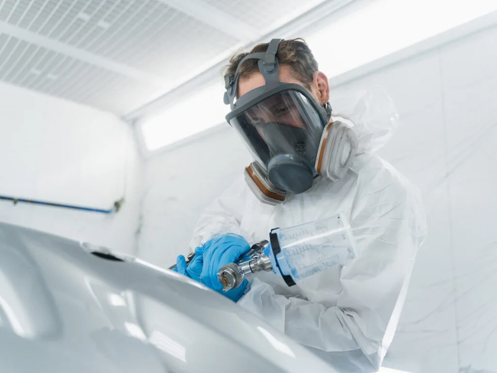 Kaltenbach Gruppe – Mit Schutzanzug, Handschuhen und Atemschutzmaske lackiert ein Techniker mit einer Spritzpistole in einem gut beleuchteten, geschlossenen Arbeitsbereich ein Autoteil von Kaltenbach Classic. – BMW, MINI, KIA, BMW Motorrad & Honda Motorrad Partner in NRW