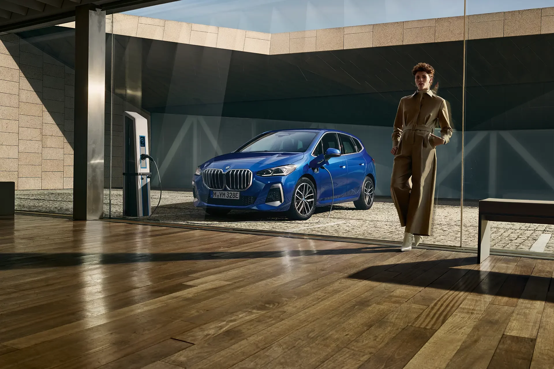 Kaltenbach Gruppe – Eine Person in einem hellbraunen Mantel geht im Innenbereich der BMW Markenwelt in der Nähe einer großen Glaswand spazieren, während draußen ein blaues BMW Auto vorfährt. Das Sonnenlicht wirft Schatten auf das moderne Gebäude und den Holzboden. – BMW, MINI, KIA, BMW Motorrad & Honda Motorrad Partner in NRW
