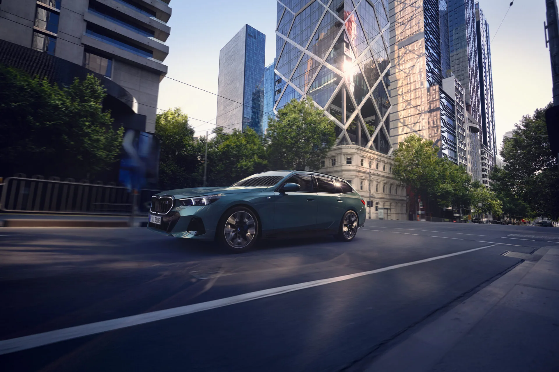 Kaltenbach Gruppe – Ein schnittiger, blauer BMW gleitet durch eine ruhige Stadtstraße, umgeben von hohen, modernen Glasgebäuden und grünen Bäumen im hellen Tageslicht - eine perfekte Szene aus der BMW Markenwelt. – BMW, MINI, KIA, BMW Motorrad & Honda Motorrad Partner in NRW