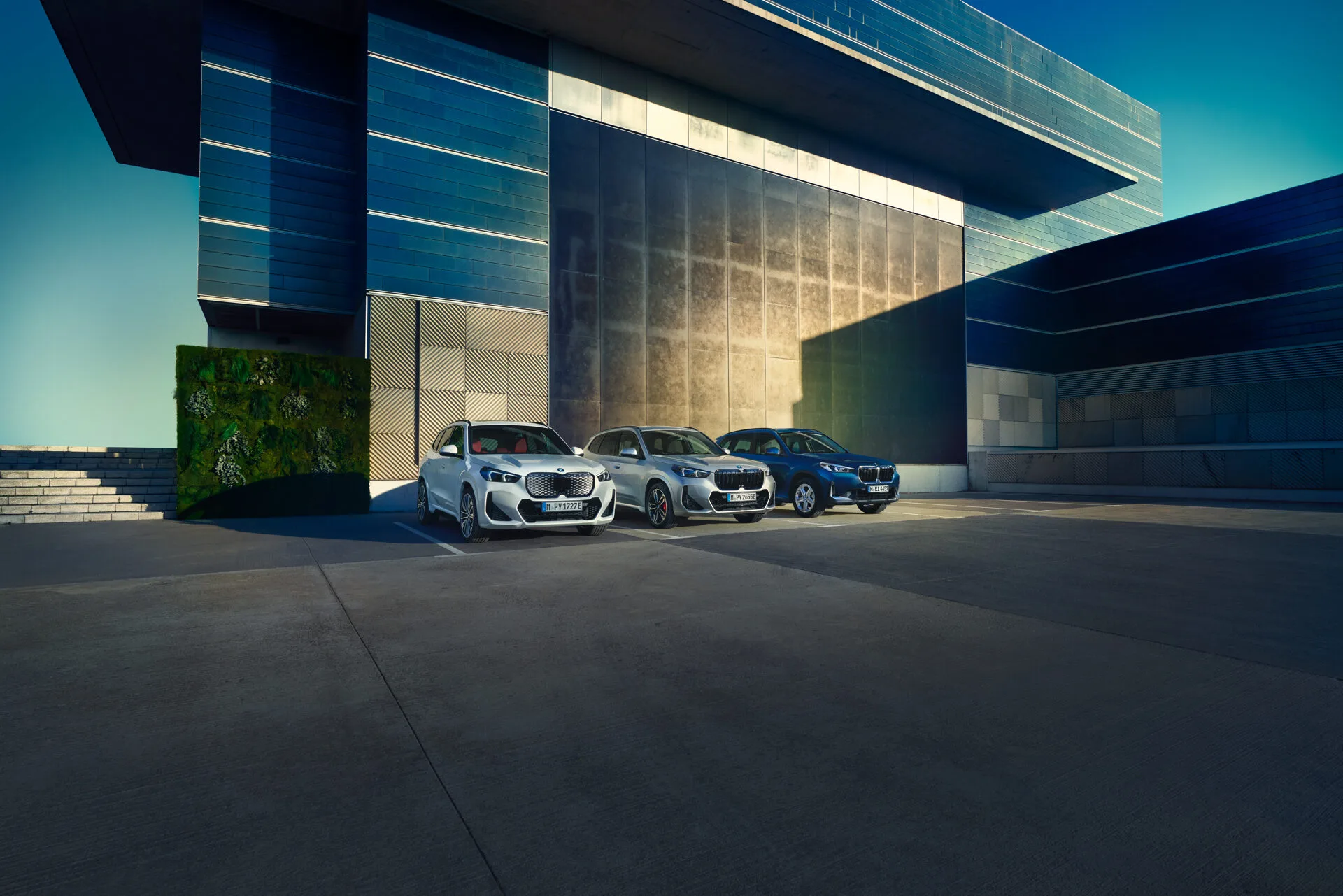 Kaltenbach Gruppe – Drei BMW Fahrzeuge sind nebeneinander vor einem modernen Gebäude mit blauen und metallischen Paneelen geparkt und zeigen den Geist der Markenwelt an einem sonnigen Tag, während sich lange Schatten über den geräumigen Betonplatz erstrecken. – BMW, MINI, KIA, BMW Motorrad & Honda Motorrad Partner in NRW