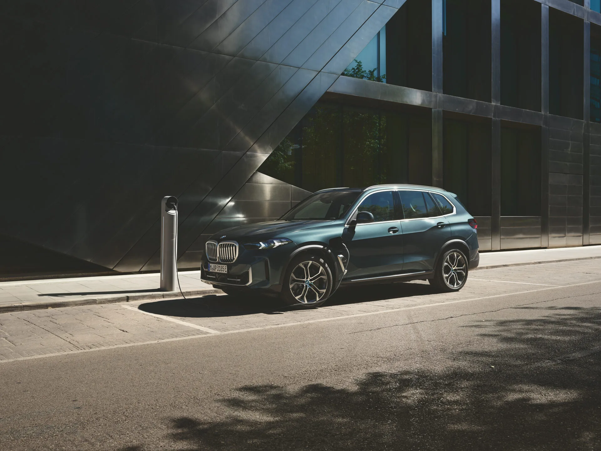 Kaltenbach Gruppe – Ein dunkelgrüner BMW Geländewagen parkt auf einer Stadtstraße neben einem modernen Gebäude, angeschlossen an eine Ladestation für Elektrofahrzeuge im hellen Tageslicht und zeigt den Innovationsgeist der BMW Markenwelt. – BMW, MINI, KIA, BMW Motorrad & Honda Motorrad Partner in NRW