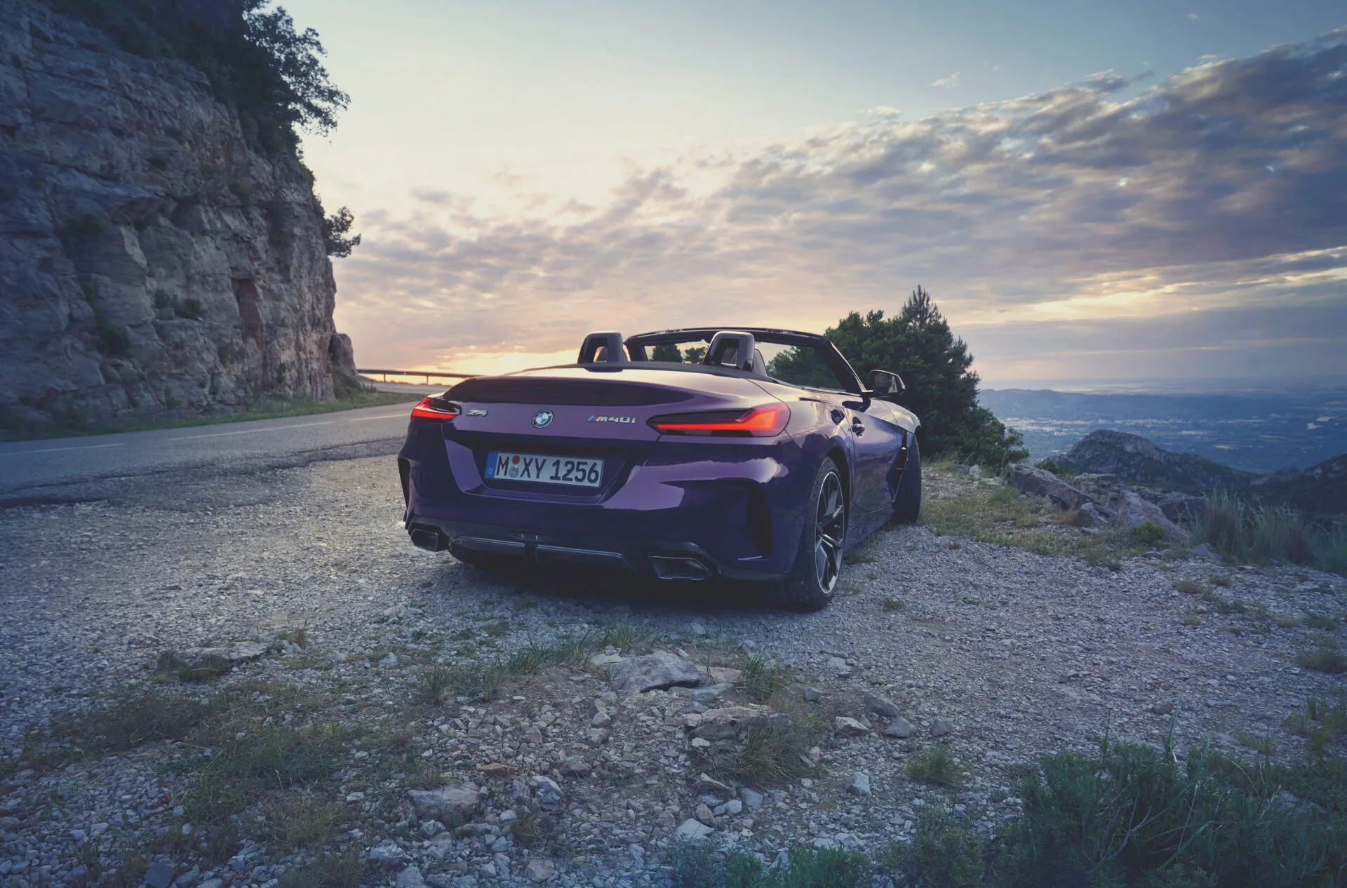 Kaltenbach Gruppe – Ein lilafarbenes BMW Z4 Cabrio, das an einem felsigen Straßenrand geparkt ist, blickt bei Sonnenuntergang auf ein malerisches Tal und zeigt die dramatischen Wolken und Klippen, die die Eleganz der BMW Markenwelt unterstreichen. – BMW, MINI, KIA, BMW Motorrad & Honda Motorrad Partner in NRW