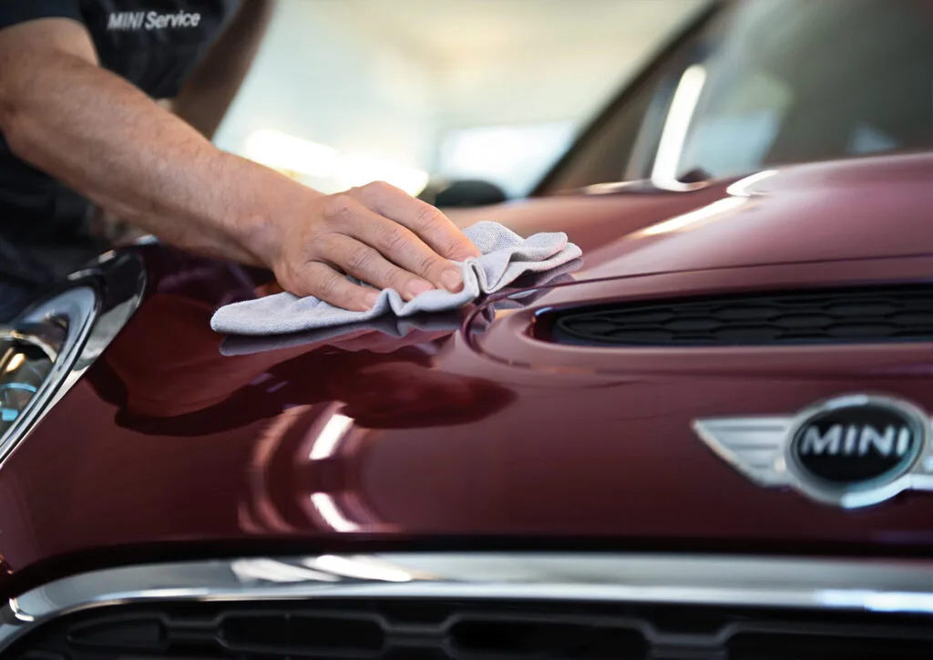 Kaltenbach Gruppe – Eine Person poliert mit einem Mikrofasertuch die glänzende kastanienbraune Motorhaube eines MINI in der Markenwelt, auf deren Vorderseite das MINI Logo zu sehen ist. Die Person trägt ein Hemd mit dem Aufdruck MINI Service auf dem Ärmel. – BMW, MINI, KIA, BMW Motorrad & Honda Motorrad Partner in NRW