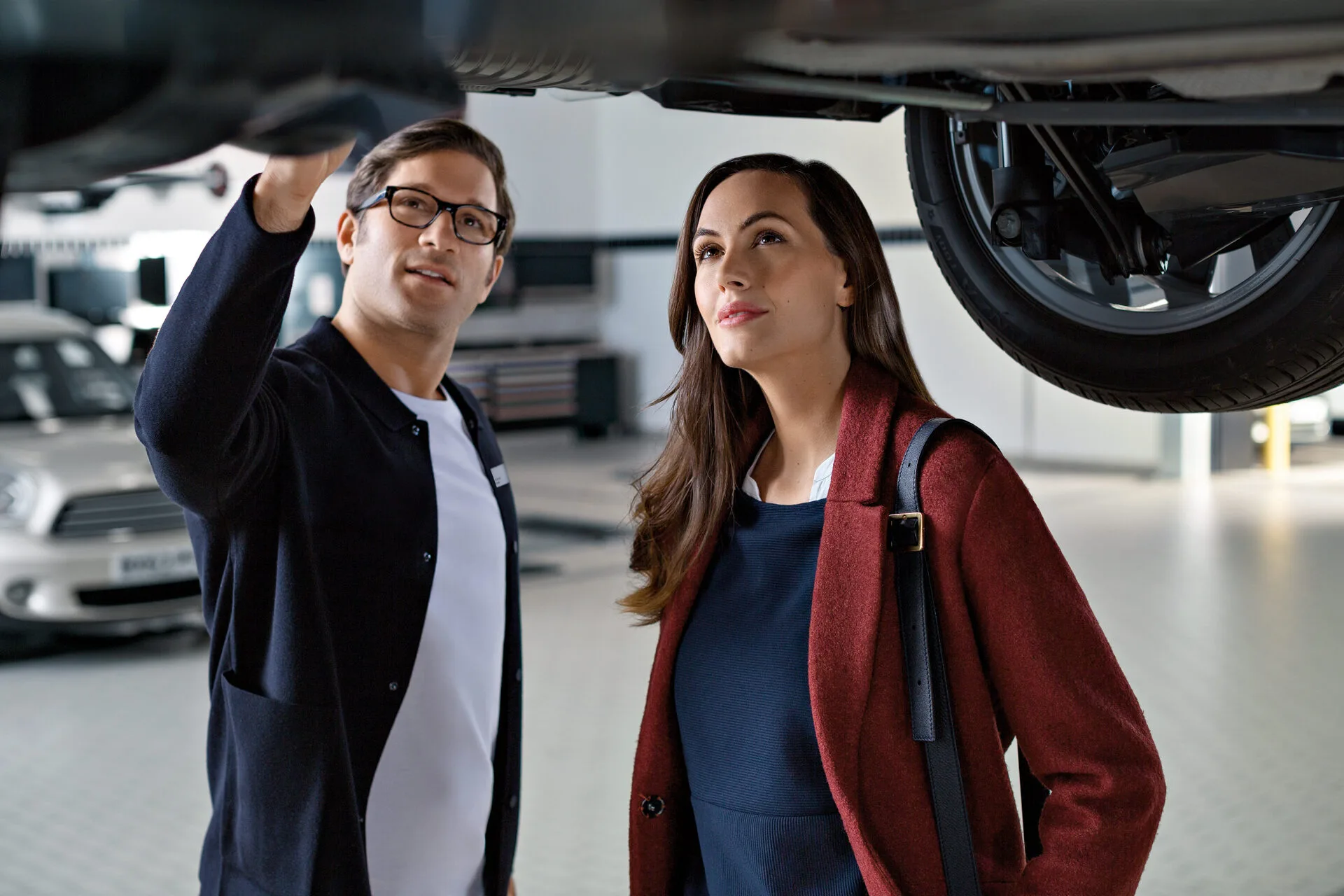Kaltenbach Gruppe – Ein Mann und eine Frau stehen in einer Autowerkstatt und schauen auf die Unterseite eines MINI. Der Mann zeigt auf etwas darüber, während die Frau aufmerksam zusieht. Beide sind in Business Casual gekleidet und erkunden gemeinsam die MINI Markenwelt. – BMW, MINI, KIA, BMW Motorrad & Honda Motorrad Partner in NRW