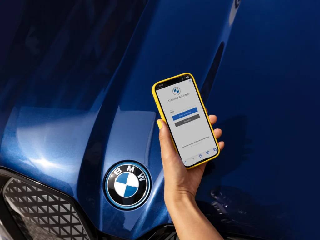 Kaltenbach Gruppe – Eine Hand hält ein Smartphone mit einem gelben Gehäuse, das einen Anmeldebildschirm für eine BMW App zeigt, über der Motorhaube eines blauen BMW Fahrzeugs, auf dem das BMW Logo zu sehen ist. – BMW, MINI, KIA, BMW Motorrad & Honda Motorrad Partner in NRW