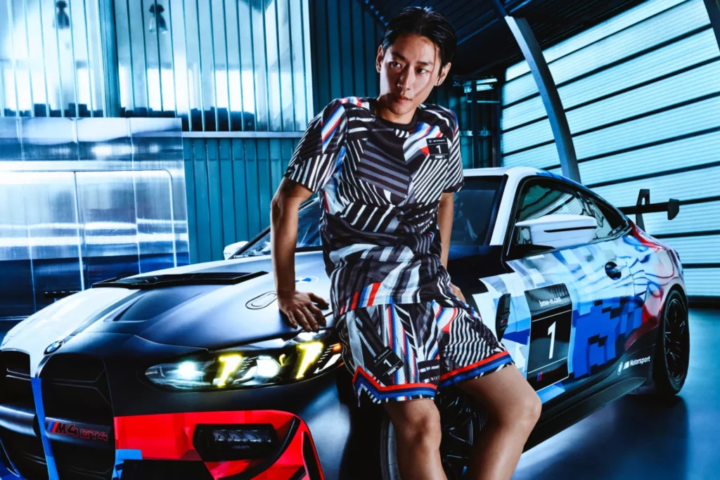 Kaltenbach Gruppe – Eine Person in einem geometrisch gemusterten Outfit sitzt auf der Motorhaube eines farbenfrohen, vom Rennsport inspirierten BMW Fahrzeugs in einer modernen Garage im Industriestil, die den Geist der BMW Markenwelt verkörpert. – BMW, MINI, KIA, BMW Motorrad & Honda Motorrad Partner in NRW