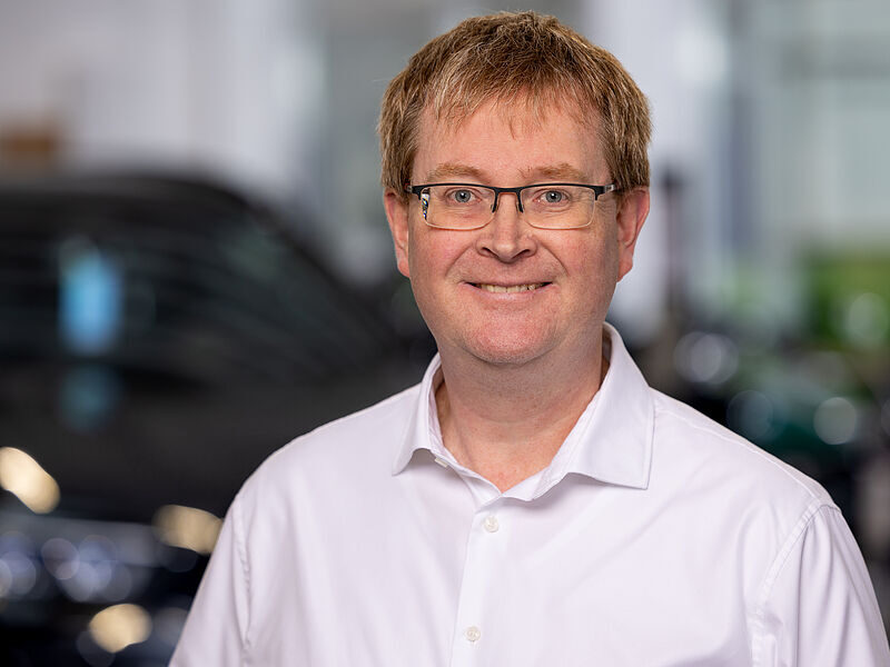 Kaltenbach Gruppe – Ein Mann mit kurzen hellbraunen Haaren und Brille, der ein weißes Button-up-Hemd trägt, lächelt in die Kamera. Der unscharfe Hintergrund deutet auf einen Innenraum in Kaltenbach Lüdenscheid hin, in dem Andeutungen eines Autos zu sehen sind. – BMW, MINI, KIA, BMW Motorrad & Honda Motorrad Partner in NRW