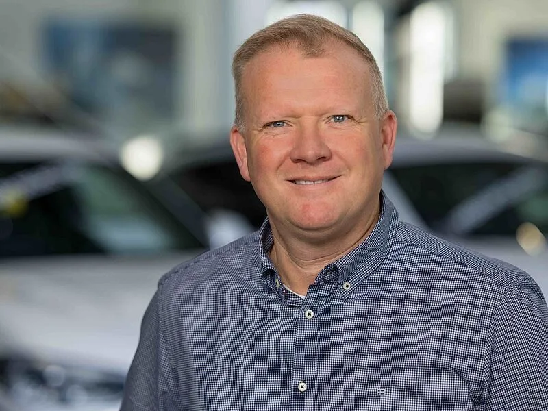 Kaltenbach Gruppe – Ein Mann mit kurzen blonden Haaren, der ein blau kariertes Button-Down-Hemd trägt, lächelt, während er in einem Haus vor verschwommenen Autos steht. Der Hintergrund ist hell und modern. – BMW, MINI, KIA, BMW Motorrad & Honda Motorrad Partner in NRW
