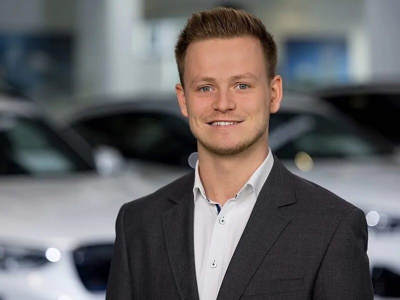 Kaltenbach Gruppe – Ein junger Mann in einem dunklen Anzug und weißem Hemd lächelt selbstbewusst, während er in Kaltenbach Lennestadt vor verschwommenen weißen Autos steht, was auf ein Autohaus oder einen Ausstellungsraum schließen lässt. – BMW, MINI, KIA, BMW Motorrad & Honda Motorrad Partner in NRW