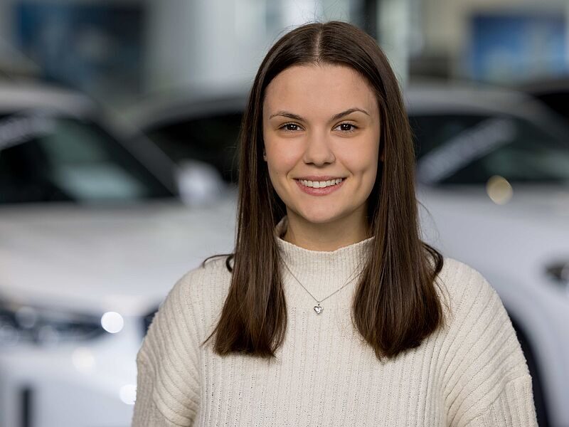 Kaltenbach Gruppe – Eine junge Frau mit langen braunen Haaren und einem weißen Pullover lächelt in die Kamera. Sie steht in einem Innenraum in Kaltenbach Lennestadt, im Hintergrund sind unscharfe weiße Autos zu sehen. – BMW, MINI, KIA, BMW Motorrad & Honda Motorrad Partner in NRW