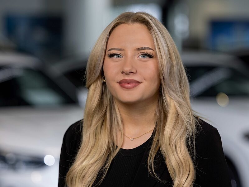 Kaltenbach Gruppe – Eine junge Frau mit langem, gewelltem blondem Haar und leichtem Make-up lächelt leicht. Sie trägt ein schwarzes Oberteil und eine zarte Halskette. Sie steht in einem Innenraum in Kaltenbach Lüdenscheid mit unscharfen Autos im Hintergrund. – BMW, MINI, KIA, BMW Motorrad & Honda Motorrad Partner in NRW
