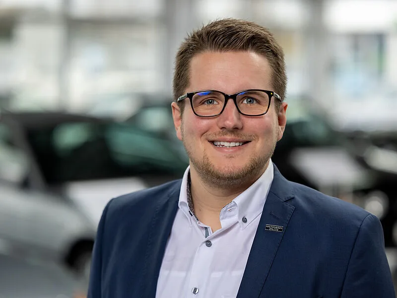 Kaltenbach Gruppe – Ein lächelnder Mann mit kurzen braunen Haaren, Brille und Gesichtsbehaarung, der eine blaue Anzugsjacke und ein weißes Hemd trägt, steht in einem Haus mit unscharfen Autos im Hintergrund. – BMW, MINI, KIA, BMW Motorrad & Honda Motorrad Partner in NRW