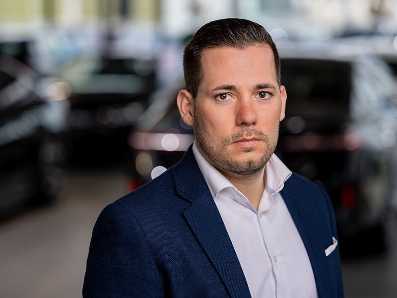Kaltenbach Gruppe – Ein Mann mit kurzen dunklen Haaren und gestutztem Bart, der eine blaue Anzugsjacke und ein weißes Hemd trägt, steht in einem Innenraum vor verschwommenen Autos und schaut ernst in die Kamera. – BMW, MINI, KIA, BMW Motorrad & Honda Motorrad Partner in NRW