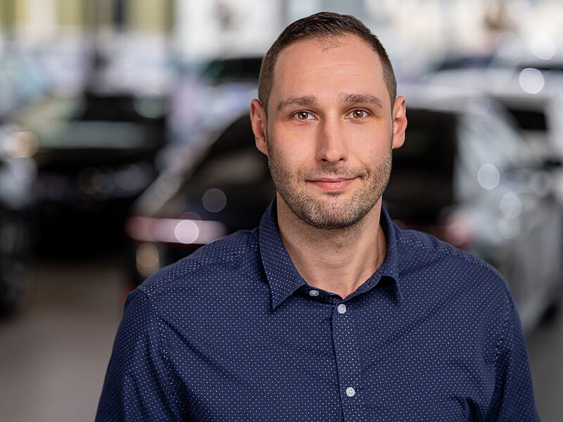 Kaltenbach Gruppe – Ein Mann mit kurzen Haaren und gestutztem Bart, der ein marineblaues Button-up-Hemd trägt, steht in einem Innenraum mit verschwommenen Autos und Lichtern im Hintergrund, blickt direkt in die Kamera und lächelt leicht. – BMW, MINI, KIA, BMW Motorrad & Honda Motorrad Partner in NRW