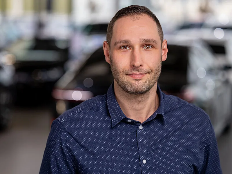 Kaltenbach Gruppe – Ein Mann mit kurzen Haaren und gestutztem Bart, der ein marineblaues Button-up-Hemd trägt, steht in einem Innenraum mit verschwommenen Autos und Lichtern im Hintergrund, blickt direkt in die Kamera und lächelt leicht. – BMW, MINI, KIA, BMW Motorrad & Honda Motorrad Partner in NRW