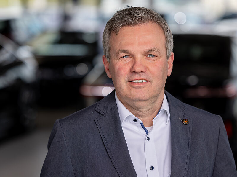 Kaltenbach Gruppe – Ein Mann mittleren Alters mit kurzen grauen Haaren trägt ein hellblaues Hemd und einen dunklen Blazer und lächelt in die Kamera. Im Hintergrund sind dunkel gefärbte Autos zu sehen, die an einen Ausstellungsraum erinnern. – BMW, MINI, KIA, BMW Motorrad & Honda Motorrad Partner in NRW