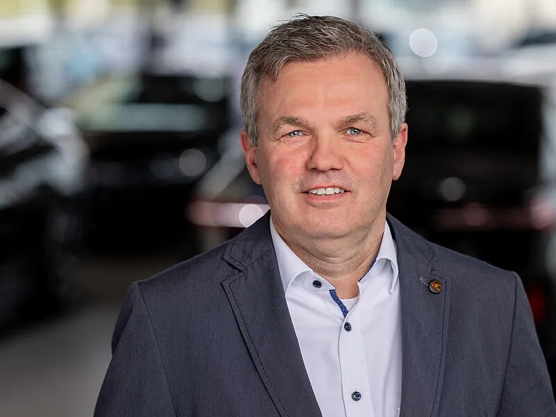 Kaltenbach Gruppe – Ein Mann mittleren Alters mit kurzen grauen Haaren trägt ein hellblaues Hemd und einen dunklen Blazer und lächelt in die Kamera. Im Hintergrund sind dunkel gefärbte Autos zu sehen, die an einen Ausstellungsraum erinnern. – BMW, MINI, KIA, BMW Motorrad & Honda Motorrad Partner in NRW