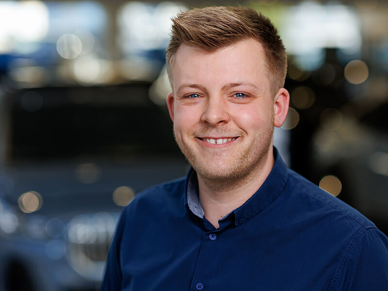 Kaltenbach Gruppe – Ein lächelnder junger Mann mit kurzen hellbraunen Haaren und gestutztem Bart, der ein dunkelblaues Button-up-Hemd trägt, steht in einem Innenraum vor einem verschwommenen Hintergrund mit einem dunklen Auto und hellen Lichtern. – BMW, MINI, KIA, BMW Motorrad & Honda Motorrad Partner in NRW
