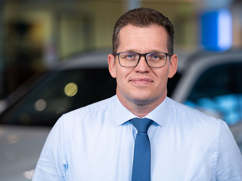 Kaltenbach Gruppe – Ein Mann, der eine Brille, ein hellblaues Hemd und eine blaue Krawatte trägt, steht in einem Innenraum vor einem verschwommenen Auto in Kaltenbach Lennestadt. Er hat kurzes braunes Haar und einen neutralen Gesichtsausdruck. – BMW, MINI, KIA, BMW Motorrad & Honda Motorrad Partner in NRW