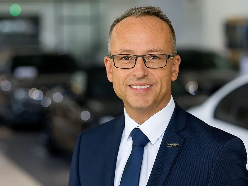 Kaltenbach Gruppe – Ein Mann mit Brille und marineblauem Anzug mit Krawatte lächelt in die Kamera im modernen Ausstellungsraum von Kaltenbach Bergisch Gladbach, mit unscharfen Autos im Hintergrund. – BMW, MINI, KIA, BMW Motorrad & Honda Motorrad Partner in NRW