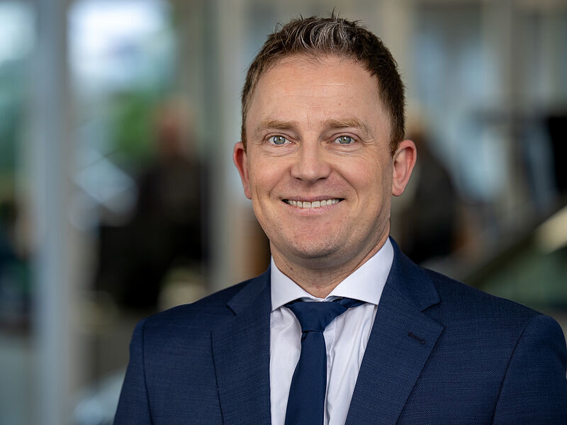 Kaltenbach Gruppe – Ein Mann in blauem Anzug und Krawatte lächelt in die Kamera in einem hell erleuchteten Innenraum, mit unscharfen Menschen und Fenstern im Hintergrund - ein professioneller Moment bei Kaltenbach Lüdenscheid. – BMW, MINI, KIA, BMW Motorrad & Honda Motorrad Partner in NRW