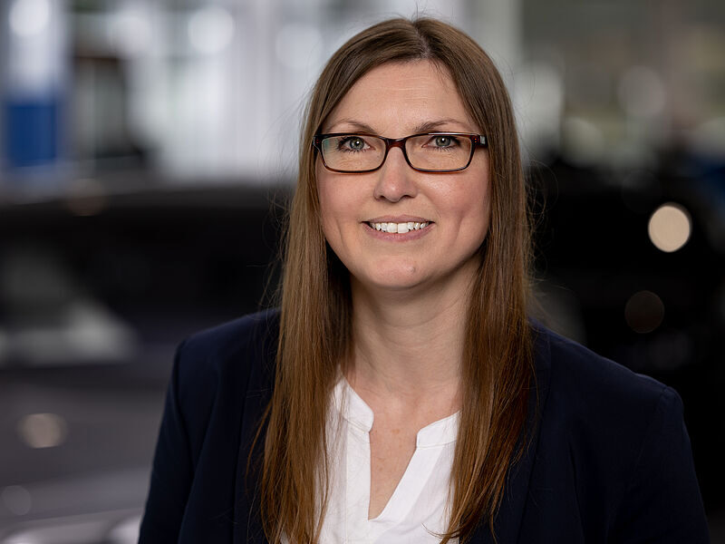 Kaltenbach Gruppe – Eine Frau mit langen glatten braunen Haaren, die eine Brille, eine weiße Bluse und einen marineblauen Blazer trägt, lächelt in die Kamera. Der Hintergrund ist unscharf und deutet auf einen Innenbereich hin, möglicherweise ein Büro oder einen Ausstellungsraum. – BMW, MINI, KIA, BMW Motorrad & Honda Motorrad Partner in NRW