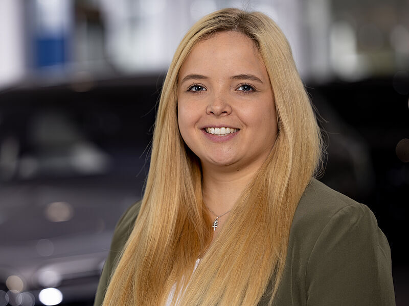 Kaltenbach Gruppe – Eine junge Frau mit langen blonden Haaren lächelt in die Kamera. Sie trägt einen grünen Blazer über einem weißen Oberteil und eine Halskette mit einem Kreuzanhänger. Der Hintergrund ist unscharf, möglicherweise ein Innenraum, in dem Autos zu sehen sind. – BMW, MINI, KIA, BMW Motorrad & Honda Motorrad Partner in NRW