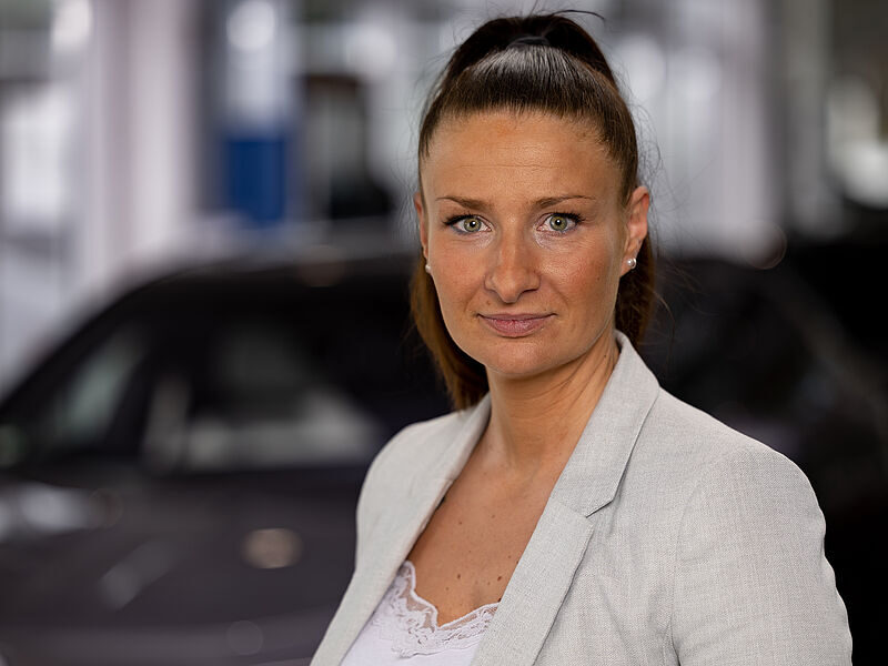 Kaltenbach Gruppe – Eine Frau mit langen braunen Haaren in einem hohen Pferdeschwanz, die einen hellgrauen Blazer und ein weißes Spitzentop trägt, steht in einem Haus in Kaltenbach Lennestadt, mit einem verschwommenen dunklen Auto im Hintergrund. Sie blickt selbstbewusst in die Kamera. – BMW, MINI, KIA, BMW Motorrad & Honda Motorrad Partner in NRW