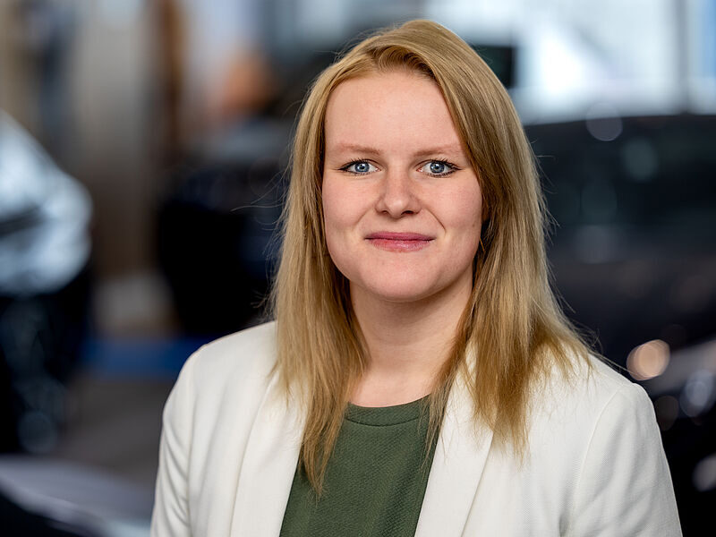 Kaltenbach Gruppe – Eine Frau mit glattem blondem Haar, die einen weißen Blazer über einem grünen Oberteil trägt, lächelt in einer unscharfen Innenaufnahme in Kaltenbach Meinerzhagen in die Kamera. – BMW, MINI, KIA, BMW Motorrad & Honda Motorrad Partner in NRW