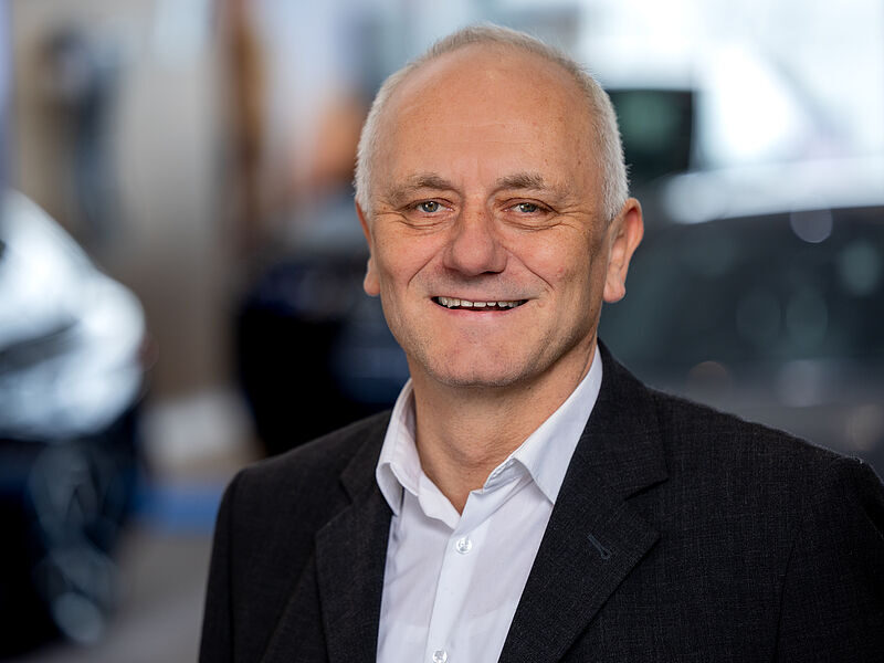 Kaltenbach Gruppe – Ein Mann mittleren Alters mit kurzen grauen Haaren, der ein weißes Hemd und einen dunklen Blazer trägt, lächelt vor einem unscharfen Innenhintergrund in die Kamera, vielleicht bei einer Veranstaltung in Kaltenbach Meinerzhagen. – BMW, MINI, KIA, BMW Motorrad & Honda Motorrad Partner in NRW