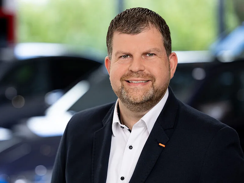 Kaltenbach Gruppe – Ein Mann mit kurzen braunen Haaren und einem Bart, der ein weißes Hemd und einen dunklen Blazer trägt, lächelt, während er in einem Innenraum in Kaltenbach Bergisch Gladbach posiert, mit unscharfen Autos und Grünflächen im Hintergrund. – BMW, MINI, KIA, BMW Motorrad & Honda Motorrad Partner in NRW